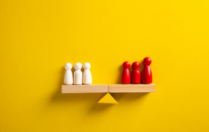 A wooden balance scale against a bright yellow background, with three white wooden figurines on the left side and three red wooden figurines on the right side. The scale is tilted, indicating an imbalance between the two groups.