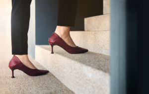 A close-up of a person's legs as they walk up a set of stairs. The person is wearing black pants and red high-heeled shoes. The focus is on the shoes and the steps, highlighting a sense of movement and progression.