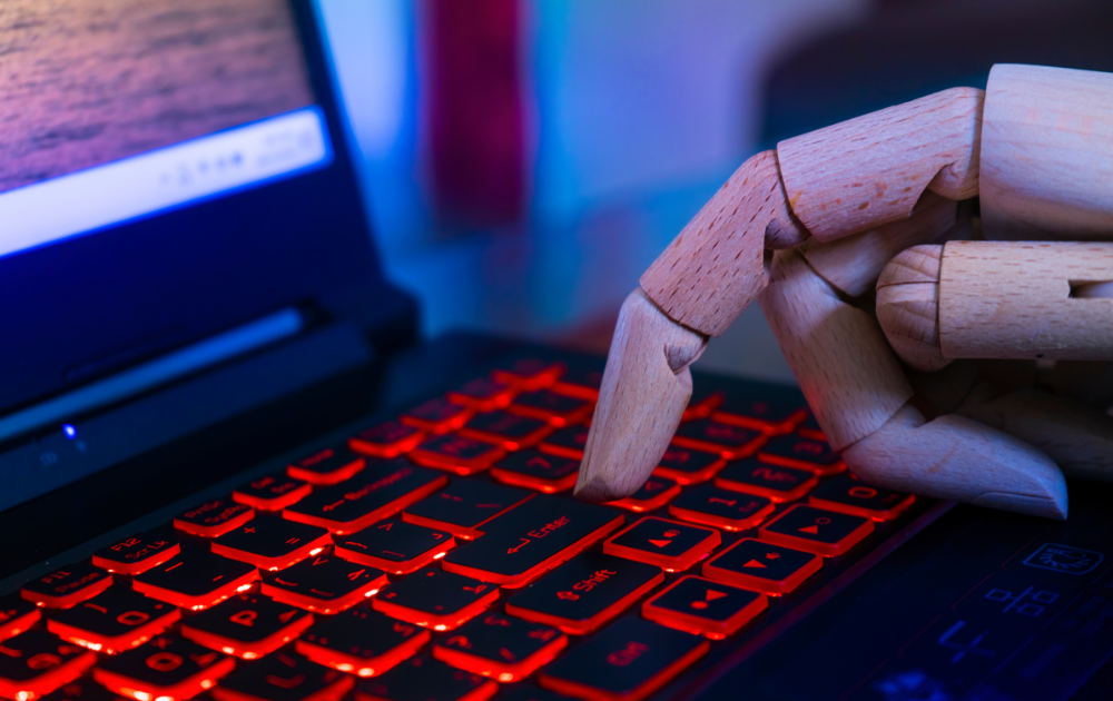 A robotic hand is seen typing on a laptop with illuminated red keys. The laptop screen displays a blurred image with warm tones, contrasting with the cool blue lighting in the background. The scene creates a futuristic and somewhat surreal atmosphere, symbolizing the integration of AI in technology.