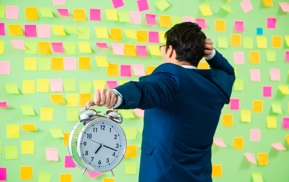 A man in a blue suit holds an alarm clock in his outstretched left hand while scratching his head with his right hand. He stands in front of a wall covered with colorful sticky notes, looking overwhelmed and confused. The sticky notes are arranged in a random pattern, creating a chaotic and cluttered background.