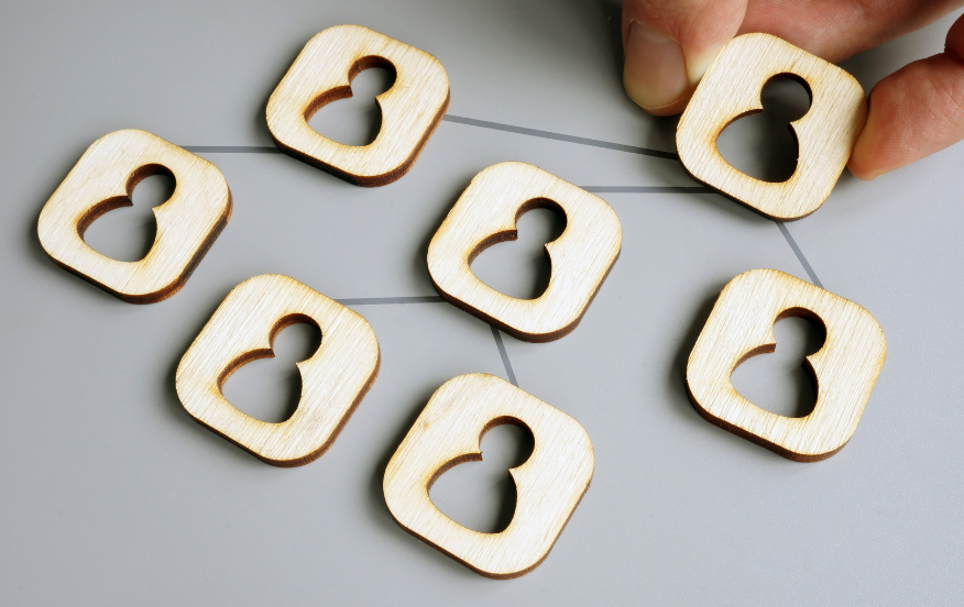 A hand arranging six wooden icons shaped like people on a gray surface, forming a network diagram. Each icon represents a team member, illustrating the concept of delegation and teamwork.