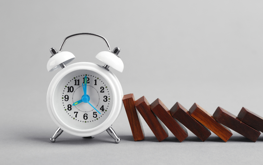 A white alarm clock with blue hands stands on a gray surface next to a row of wooden dominoes that are beginning to topple over. The scene symbolizes the concept of time and the chain reaction of events, illustrating the importance of timing and its impact.