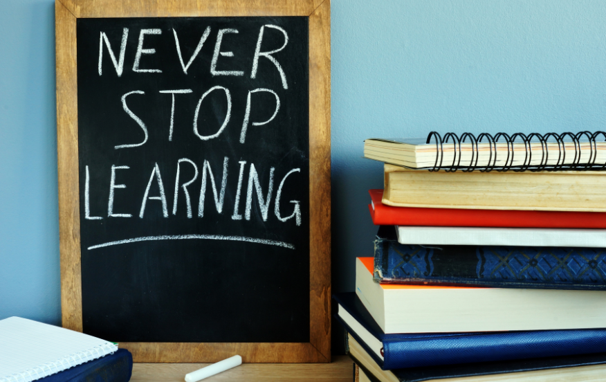 A small chalkboard with a wooden frame displays the handwritten message "Never Stop Learning" in white chalk. The chalkboard is placed next to a stack of books, notebooks, and spiral-bound journals on a wooden surface, against a light blue wall. A piece of chalk rests on the surface in front of the chalkboard, emphasizing the theme of continuous education and personal growth.