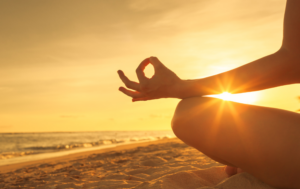 A person meditating on a beach at sunset, with the sun glowing brightly behind their hand, which is positioned in a classic meditation pose. The golden light of the setting sun creates a serene and peaceful atmosphere.