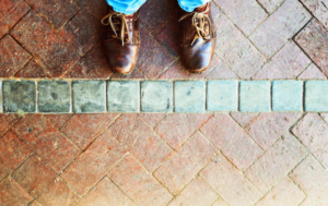 A pair of feet in brown leather shoes standing on a brick pathway with a line of gray square tiles in the middle. The person is wearing light blue jeans, and the bricks are arranged in a herringbone pattern, creating a visually interesting texture.