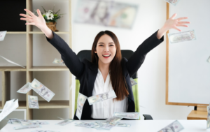 A smiling woman in a business suit sits at a desk in an office, joyfully throwing money into the air. Banknotes are floating around her, creating a celebratory and prosperous atmosphere. The scene conveys a sense of financial success and abundance. In the background, there are shelves with decorative items and a whiteboard.