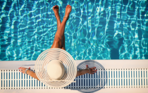 The image shows a person sitting poolside, viewed from above, with their legs crossed and feet hovering over the water. The person is wearing a large, wide-brimmed sun hat, shading their face. They are resting their arms on the pool's edge, with the clear blue water reflecting sunlight in mesmerizing patterns. The scene evokes a sense of relaxation and tranquility, perfect for a sunny day by the pool.