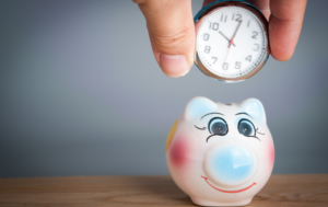 A hand is placing a small clock into the slot of a smiling piggy bank, symbolizing the concept of saving time. The piggy bank has a cheerful face with rosy cheeks, and the clock represents time management or the value of time. The background is neutral, highlighting the focus on the piggy bank and clock