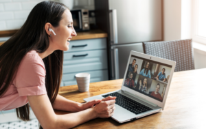 A smiling woman wearing wireless earbuds sits at a kitchen table, engaged in a video call on her laptop. Several participants are visible on the screen. She appears happy and relaxed, interacting with the group in a casual, homey environment. A cup sits on the table beside her as she leans forward, listening attentively.