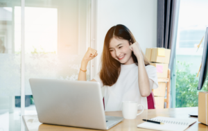 A young woman celebrates in front of her laptop, raising her fists in excitement. She smiles joyfully as she sits at a desk, clearly pleased with something on the screen. A coffee mug and notebook are on the table, and cardboard boxes are stacked behind her, suggesting a home office or entrepreneurial workspace. The sunlight streams in through a large window in the background.