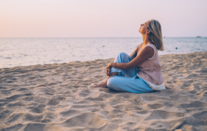 The image shows a woman sitting on the sand at the beach, facing the ocean during what appears to be a sunset or early evening. She has her knees drawn up and arms wrapped around them, looking upwards with her eyes closed, seemingly enjoying a peaceful moment. The sky has soft, pastel hues, and the calm sea stretches out in the background, creating a serene and relaxing atmosphere.