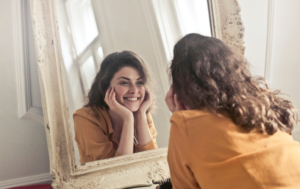 The image shows a woman in a mustard-yellow shirt smiling at her reflection in a large, vintage-style mirror. She has curly, dark brown hair and is resting her face in her hands while leaning slightly forward toward the mirror. The atmosphere appears warm and bright, creating a cheerful, uplifting mood as she gazes at herself with joy.