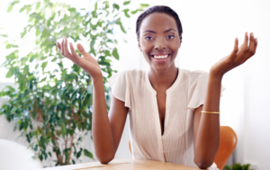 A confident woman with dark hair pulled back is smiling brightly while sitting at a table. She is wearing a light beige blouse and gesturing with both hands, palms up in an open and welcoming manner. In the background, there are green plants and bright natural light, creating a fresh and inviting atmosphere.