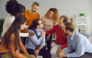 A group of diverse people gathers around a man seated in the center, offering support by placing their hands on his shoulders. The man, wearing glasses and a blue shirt, looks up at them, engaged in conversation. The group consists of men and women, dressed casually, and their body language conveys care and empathy. They are in a well-lit room with modern decor, including shelves and plants in the background.