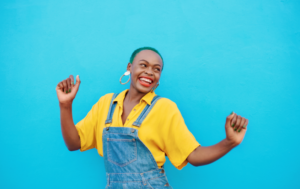 A joyful woman with short green hair exudes confidence as she smiles brightly, wearing bold red lipstick. Dressed in a vibrant yellow shirt and denim overalls, she poses energetically against a bright blue wall, raising her arms in celebration. Her radiant expression and confident posture convey a carefree and empowered attitude, full of self-assurance and positivity.