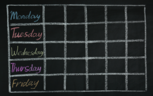 The image shows a blank chalkboard-style weekly schedule grid. The weekdays—Monday, Tuesday, Wednesday, Thursday, and Friday—are written in colorful chalk (blue, red, green, pink, and yellow, respectively) on the left side, with several empty boxes next to each day. This design provides space for adding tasks or appointments for each weekday.