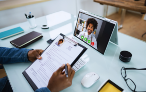 The image shows a person conducting a virtual interview, holding a resume on a clipboard while looking at a laptop or tablet screen displaying a video call. The interviewee, smiling and professionally dressed, is visible on the screen. The desk is clean and organized, with a notebook, mouse, and other office supplies, representing a modern, remote hiring process. This image conveys the idea of online interviews or virtual assistant recruitment.