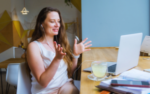 The image shows a young woman sitting at a wooden table in a casual setting, engaging in a video call on her laptop. She is smiling and gesturing with her hands while wearing earphones, indicating an interactive and friendly conversation. A glass of lemon water and a few notebooks are on the table beside her, adding to the relaxed yet productive atmosphere. This image represents virtual meetings or online coaching sessions.