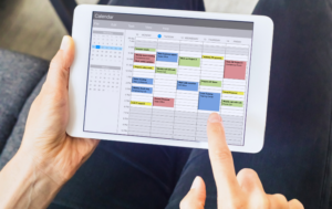 The image shows a person holding a tablet with an open calendar app. The screen displays a detailed schedule for the week, with various appointments and tasks organized by time slots in different colors. The individual is pointing to a specific task on Tuesday, indicating interaction with the schedule. This visual represents digital time management, productivity tools, or organizing a busy workweek.